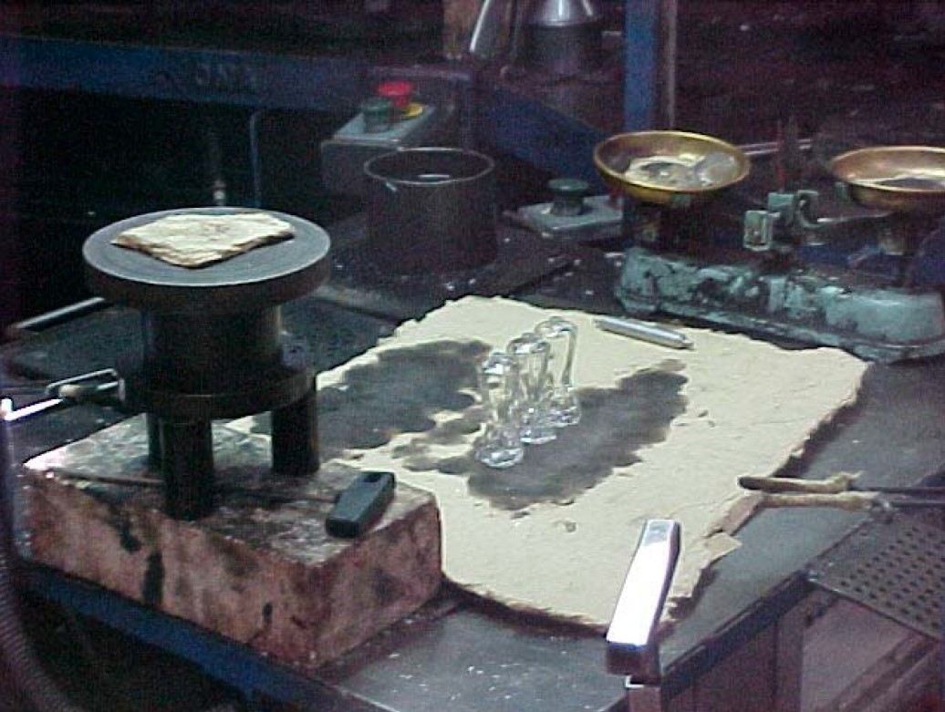 A la imatge veiem un taller artesanal antic amb un cartró d'amiant sobre la taula, que serveix de base per dipositar-hi unes peces de vidre que s'acaben de fabricar.