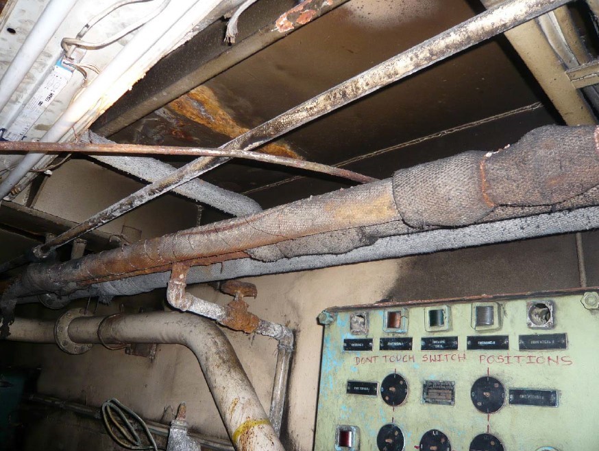 A la imatge veiem uns feltres d'amiant teixits embolcallant tubs d'instal·lacions que discorren per un sostre i sobre un quadre elèctric antic.